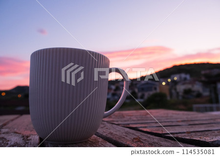 As the sun sets, a coffee cup is placed on a wooden table against a backdrop of the colorful sky. The serene landscape is enhanced by the simple beauty of the tableware 114535081