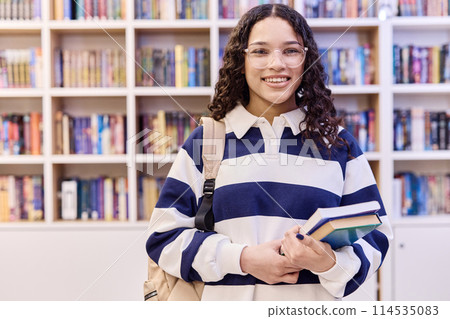 Waist up portrait of smiling teenage girl wearing glasses and looking at camera standing in school library with books and backpack copy space 114535083