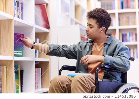 Side view portrait of young teenage boy with disability using wheelchair in library and choosing books on shelf 114535090