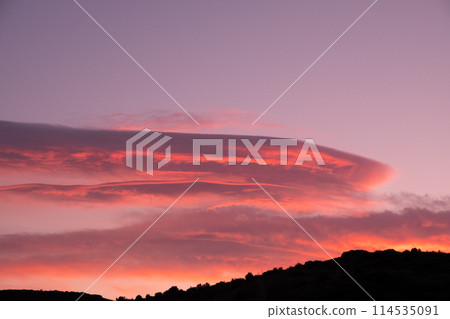 A picturesque sunset sky with a few cumulus clouds, creating a stunning atmosphere with shades of orange and red. The natural landscape is bathed in the afterglow of dusk 114535091
