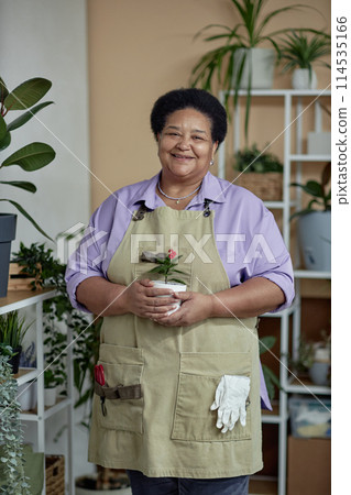 Vertical portrait of kind Black senior woman holding green plant and smiling at camera while enjoying gardening Vertical portrait of kind Black senior woman holding green plant and smiling at camera while enjoying gardening 114535166