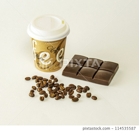 Paper cup with tiles of dark chocolate and coffee beans scattered on a gray background 114535587