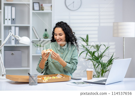 A young Hispanic woman savors a slice of pizza while sitting at her desk in a bright, well-organized office, reflecting a moment of joy amidst work. A young Hispanic woman savors a slice of pizza while sitting at her desk in a bright, well-organized office, reflecting a moment of joy amidst work. 114537656