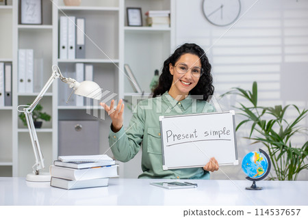 Smiling Hispanic woman teacher holding a 'Present simple' sign while explaining English grammar in a virtual classroom setup. Smiling Hispanic woman teacher holding a 'Present simple' sign while explaining English grammar in a virtual classroom setup. 114537657