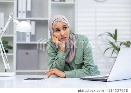 A young Middle Eastern woman wearing a hijab sits at her desk in an office, appearing pensive and thoughtful with a laptop and notebook in front of her. A young Middle Eastern woman wearing a hijab sits at her desk in an office, appearing pensive and thoughtful with a laptop and notebook in front of her. 114537658