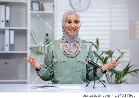 Engaging young Muslim woman speaking energetically on a podcast, addressing the camera in a well-lit, stylish office setting. 114537665