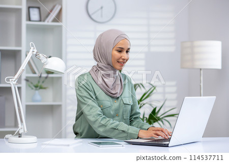 A Muslim woman in a hijab works diligently on her laptop at a well-organized office desk, surrounded by light and modern decor. A Muslim woman in a hijab works diligently on her laptop at a well-organized office desk, surrounded by light and modern decor. 114537711