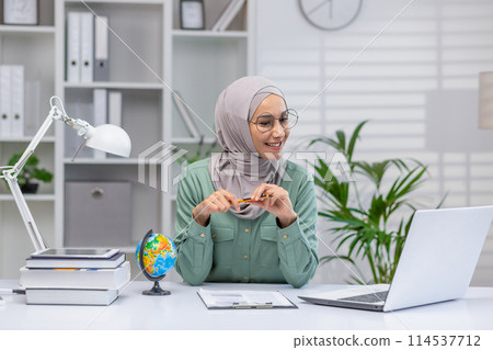 A cheerful Muslim woman wearing a hijab engaged in online teaching, using a laptop and pointing at a small globe, surrounded by books in a well-organized office. 114537712