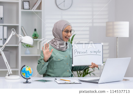 Smiling Muslim educator waves while teaching English online, displaying 'Present simple' on a whiteboard in a light-filled, organized office space. 114537713