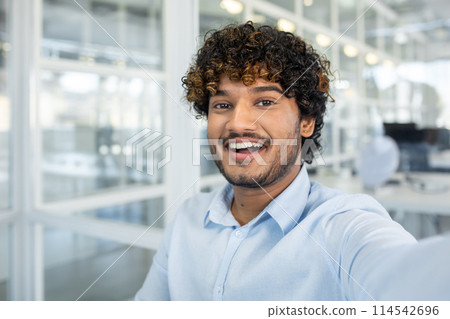 Cheerful young man snapping a selfie in a bright, modern office space, showcasing joy and a professional atmosphere. 114542696