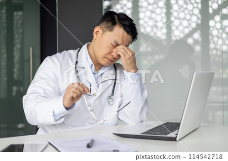 Asian mature doctor looking stressed while sitting at desk with laptop and holding glasses, conveying stress and fatigue. Asian mature doctor looking stressed while sitting at desk with laptop and holding glasses, conveying stress and fatigue. 114542718
