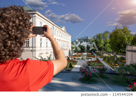 Salzburg, Austria, August 15, 2022. Close-up on a young man taking a daytime shot of the Mirabell Palace gardens. People visit them and explore. The Hohensalzburg fortress in the background. Salzburg, Austria, August 15, 2022. Close-up on a young man taking a daytime shot of the Mirabell Palace gardens. People visit them and explore. The Hohensalzburg fortress in the background. 114544104