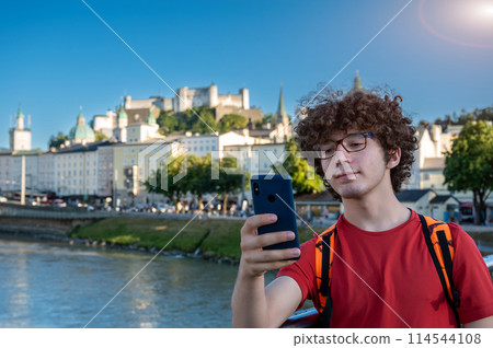 Salzburg, Austria, August 15, 2022. Nice daytime shot with a young Caucasian man taking a selfie from the Padlock Bridge with the old town dominated by the fort in the background. 114544108