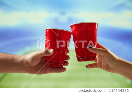 Hands holding red mugs with beer and clicking with blurred sport open air stadium on background 114544225