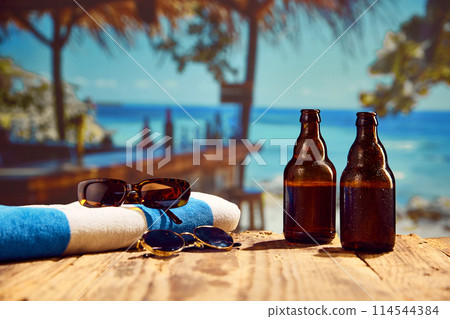 Beachside relaxation. Sunglasses, towel and cold beer bottles standing on a wooden table on beach bar with ocean background 114544384