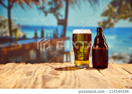 Beachside chills. Frosty beer glass and bottle wooden table, with ocean background on warm day. Beach bar 114544386