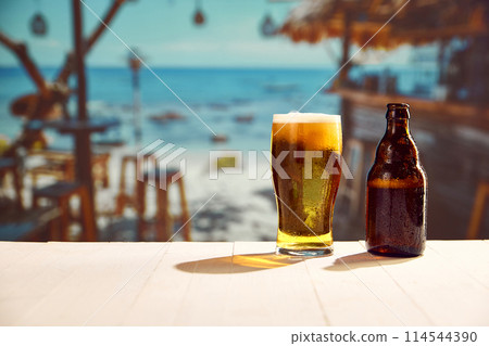 Glass with lager foamy chill beer and beer bottles standing on wooden table at local bar on beach. Sea background 114544390