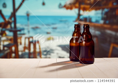 Two beer bottles standing on wooden table with beautiful ocean view, beach with resting zone, cafes and bars Two beer bottles standing on wooden table with beautiful ocean view, beach with resting zone, cafes and bars 114544391