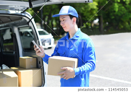 A male delivery driver holding a package and looking at his smartphone 114545053