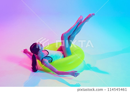 Woman in retro bikini enjoys her travelling like swimming in beach in yellow float in neon light against vibrant gradient background. Woman in retro bikini enjoys her travelling like swimming in beach in yellow float in neon light against vibrant gradient background. 114545461