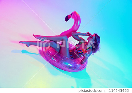 Woman lounging in pink flamingo float, drinking cocktail in neon light against vibrant gradient background. Pool party vibe Woman lounging in pink flamingo float, drinking cocktail in neon light against vibrant gradient background. Pool party vibe 114545463