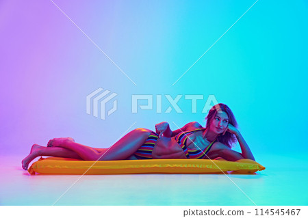 Young tender woman lying on air mattress and relaxing like swimming in ocean in neon light against vibrant gradient background. 114545467