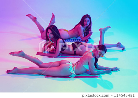 Three young, fit women lounge in style swimwear in neon light against vibrant gradient background. Summer chic. Three young, fit women lounge in style swimwear in neon light against vibrant gradient background. Summer chic. 114545481