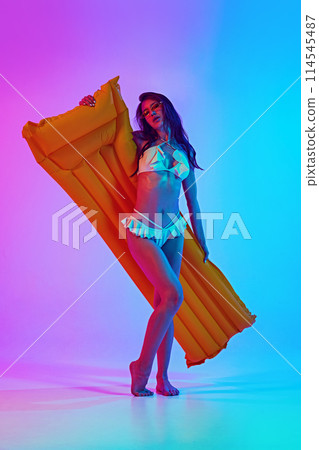Young attractive, fit woman in stylish white swimwear and sunglasses posing with air mattress in neon light against vibrant gradient background. 114545487