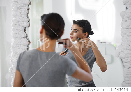 A young woman massages her face with gouache while looking in the mirror. A young woman massages her face with gouache while looking in the mirror. 114545614