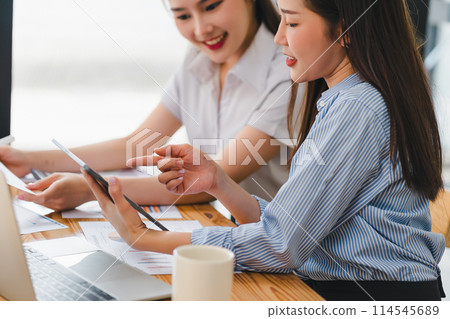 Two engaged businesswomen using a tablet and discussing work with focus and interest at an office desk. Two engaged businesswomen using a tablet and discussing work with focus and interest at an office desk. 114545689