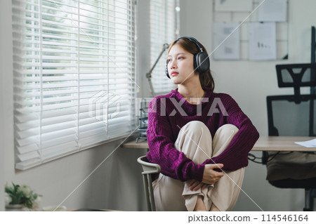 A young woman with headphones sits in a chair, listening intently, focused, and engaged in her music or podcast, feeling sad. 114546164