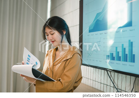 The female Chief Analyst is conducting a meeting presentation for a team of economists. The projector screen shows graphs, product sales, revenue growth strategies, and e-commerce analysis. 114546350