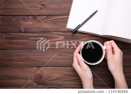 Female hand holding cup of coffee on brown wooden background. Notebook and cup of coffee on brown wooden background 114546846