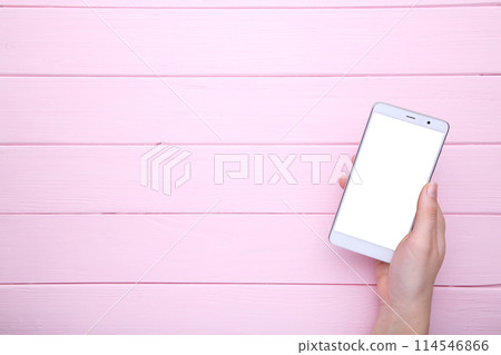 Female hands holding mobile phone with blank screen on pink wood background. Smartphone on wood table. Female hands holding mobile phone with blank screen on pink wood background. Smartphone on wood table. 114546866
