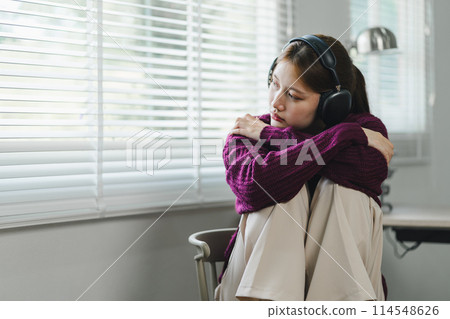 A young woman with headphones sits in a chair, listening intently, focused, and engaged in her music or podcast, feeling sad. 114548626