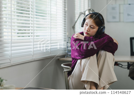 A young woman with headphones sits in a chair, listening intently, focused, and engaged in her music or podcast, feeling sad. 114548627