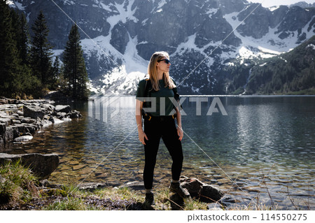 Blonde woman with backpack standing at full height by lake, mountains and snow in backdrop Blonde woman with backpack standing at full height by lake, mountains and snow in backdrop 114550275