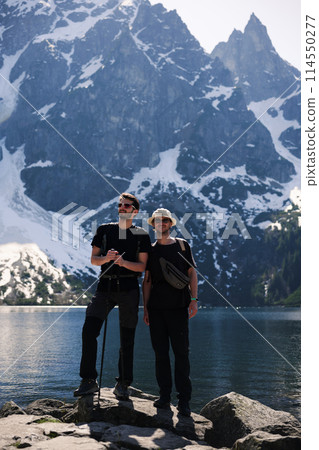 Two men on rock by lake, mountains in background, enjoying natural landscape Two men on rock by lake, mountains in background, enjoying natural landscape 114550277