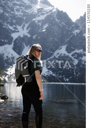 Blonde woman with backpack by lake, mountains and snow in backdrop 114550280