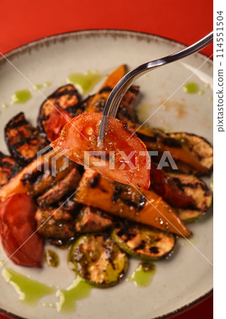 Fork with charred tomato slice over plate of grilled vegetables 114551504