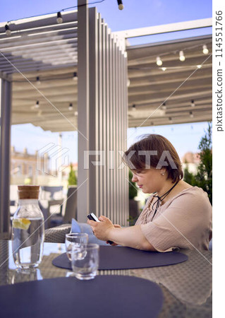 plus size woman in peach fuzz dress chooses a dish in a restaurant via QR code in smartphone, water with lemon and glasses in front of her plus size woman in peach fuzz dress chooses a dish in a restaurant via QR code in smartphone, water with lemon and glasses in front of her 114551766