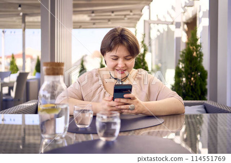 plus size woman in peach fuzz dress chooses a dish in a restaurant via QR code in smartphone, water with lemon and glasses in front of her 114551769