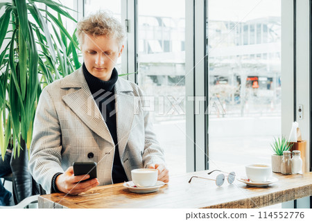 Portrait of thoughtful handsome young stylish man in checked coat using phone while drinking coffee in modern cafe shop . Cold season elegant fashion stylish outfit trend. Retro, Old money aesthetic. 114552776