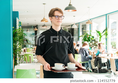 Portrait of young smiling affable waiter carrying on tray with coffee cups to client table in cafe. Hospitality service job. Occupation and work concept. Part-time work for students. Selective focus. 114552779