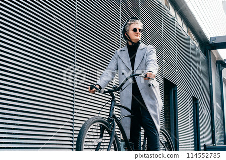 Stylish male in coat, sunglasses and protective helmet riding on retro bicycle near gray urban wall. Neutral carbon footprint transportation. Green eco friendly mobility sustainable transport. 114552785