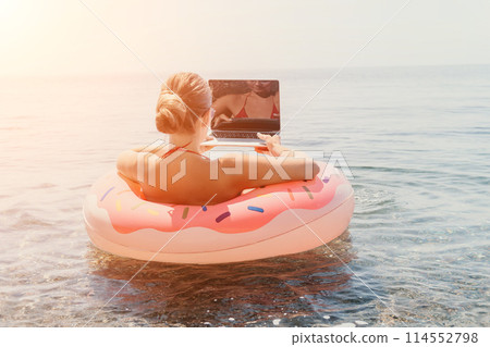 Woman freelancer works on laptop swimming in sea on pink inflatable ring. Happy tourist floating on inflatable donut and working on laptop computer in calm ocean. Freelance, remote working anywhere Woman freelancer works on laptop swimming in sea on pink inflatable ring. Happy tourist floating on inflatable donut and working on laptop computer in calm ocean. Freelance, remote working anywhere 114552798