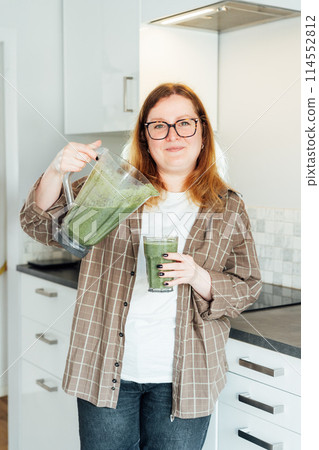 Smiling plus size young woman with just made glass of detox shake, green smoothie in the kitchen. Healthy dieting, eating, cooking, slimming. Natural weight loss program. Vegan, vegetarian diet. Smiling plus size young woman with just made glass of detox shake, green smoothie in the kitchen. Healthy dieting, eating, cooking, slimming. Natural weight loss program. Vegan, vegetarian diet. 114552812