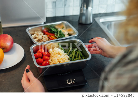 Healthy diet plan for weight loss, daily ready meal menu. Close up Woman weighing lunch box cooked in advance,ready to eat on kitchen scale. Balanced portion with healthy dish. Pre-cooking concept Healthy diet plan for weight loss, daily ready meal menu. Close up Woman weighing lunch box cooked in advance,ready to eat on kitchen scale. Balanced portion with healthy dish. Pre-cooking concept 114552845