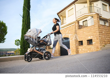 Happy woman performing morning jog and pushing a baby pram, enjoying her active lifestyle during maternity leave. People. Motherhood and babyhood concept Happy woman performing morning jog and pushing a baby pram, enjoying her active lifestyle during maternity leave. People. Motherhood and babyhood concept 114553210