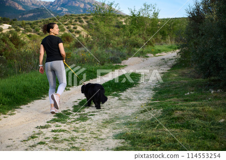 Full size shot of a happy woman smiling looking away, walking her dog in mountains nature outdoors. Rear view 114553254